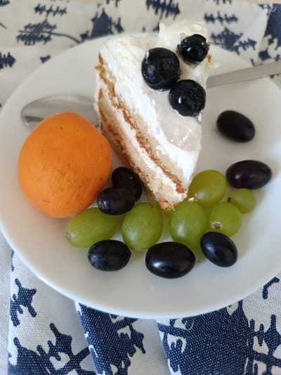 A slice of layered cake, topped with white frosting and three dark blueberries, sits on a white plate. To the left of the cake is a single orange fruit, possibly an apricot. Surrounding the cake and fruit are several green and dark purple grapes. The plate is placed on a white and navy blue patterned cloth. A silver spoon is partially visible to the left of the plate. The scene is brightly lit, suggesting an indoor setting. There are no people or animals visible in the image. The image does not provide any information about its location in Olsztyn, Poland.