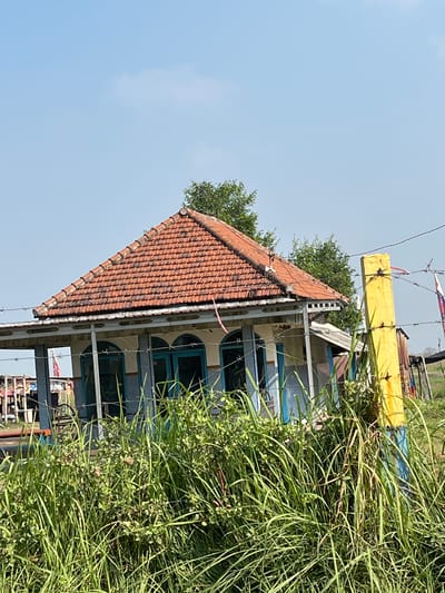 The image captures a scene in Gempolsari, Indonesia, featuring a small, weathered building with a red-tiled roof and blue-trimmed windows. The building appears to be situated in a rural or semi-rural environment, with tall green grass and vegetation in the foreground. A yellow post, possibly part of a fence, stands prominently on the right side of the frame, with barbed wire strung across it and around the scene. The sky is clear and blue, indicating daytime. The building's architecture suggests a utilitarian purpose, possibly a dwelling or a small community structure. The overall impression is one of simplicity and rural life, with elements of weather-worn structures and natural growth. There are no people visible in the image.