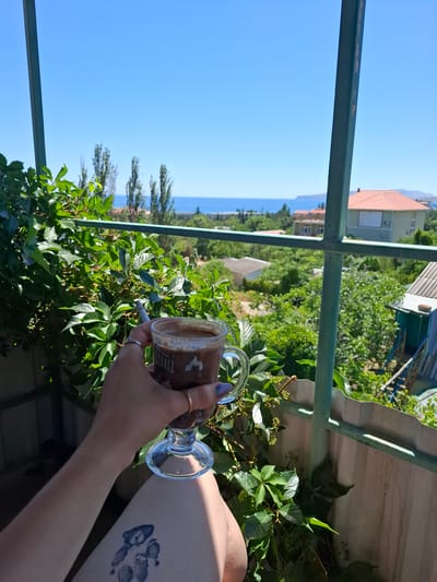 A person's hand holds a glass of coffee with a cigarette in it, while resting their arm on a railing. The person has a tattoo of paw prints on their arm and is wearing rings. The scene is outdoors, with lush green foliage in the foreground and a view of the sea and houses in the background. The sky is clear and blue, indicating a sunny day. The lighting suggests it is daytime. The overall mood is relaxed and perhaps contemplative, with the coffee and cigarette implying a moment of leisure. The visible text on the coffee cup is unclear. The location is identified as Shchebetovskyi Settlement Council, Ukraine.