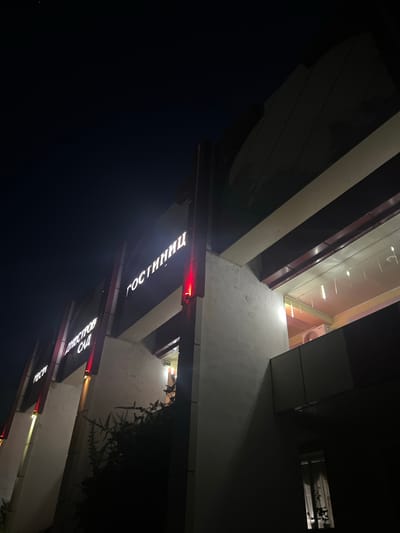 The image shows the exterior of a building at night, illuminated by its own signage and some interior lighting. The building appears to be a hotel, as indicated by the Cyrillic text "ГОСТИНИЦА" (Gostinitsa), meaning "Hotel," prominently displayed in illuminated letters on the facade. Below this, another sign reads "ДНЕСТРОВСКИЙ САД" (Dnestrovskiy Sad), which translates to "Dniester Garden." There are also illuminated letters that read "РЕСТИ" (Resti), likely a shortened version of "ресторан" (restoran), meaning "restaurant."
The architecture features a modern design with angled surfaces and a combination of light-colored concrete or stucco walls and darker paneling. Red and yellow lights are integrated into the building's structure, highlighting architectural features. The sky above is dark, suggesting it is nighttime. Interior lighting is visible through a large window on the upper level, revealing what appears to be a ceiling with hanging lights. An air conditioning unit is visible on the exterior of the building on the upper level. A dark, leafy shrub or tree is growing in front of the lower portion of the building. The overall scene is dimly lit, with the building's signage being the primary source of light. The location is identified as Dubăsari, Moldova. There are no people visible in the image.