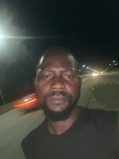 The image shows a close-up of a man's face at night. He is looking towards the camera with a serious expression. The man has dark skin, a beard, and is wearing a black t-shirt. In the background, there is a road with lights from vehicles and distant buildings, suggesting an urban or semi-urban setting. The lighting is dim, consistent with nighttime. The image appears to be taken outdoors, possibly from a moving vehicle, given the slight blur in the background and the streaking lights. There is no visible text in the image. The location context is Immigration Quarters, Nigeria.