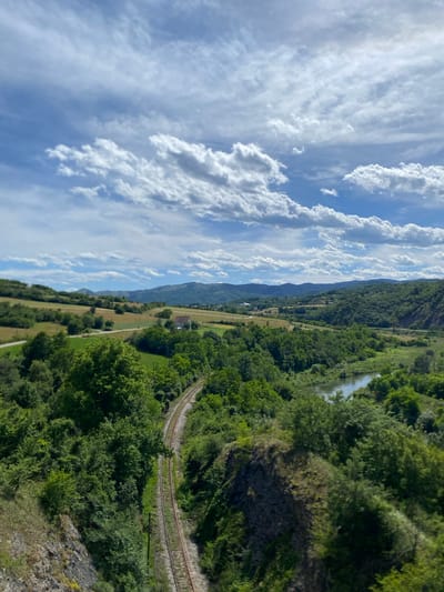 This is a high-angle, wide shot of a verdant landscape under a partly cloudy sky. In the foreground, a rocky outcrop is covered in greenery, and a set of train tracks curves away into the distance. To the right of the tracks, a calm river flows through a valley. The middle ground features rolling hills covered in lush green trees, interspersed with patches of cultivated fields. A small white building with a red roof is visible on one of the hillsides. In the background, a range of larger, blue-hued mountains stretches across the horizon, disappearing into the haze. The sky is a bright blue, filled with scattered, puffy white clouds that create a sense of depth and texture. The scene depicts a tranquil rural environment, likely in the vicinity of Raska Municipality, Serbia, characterized by its natural beauty and signs of human presence in the form of agricultural land and infrastructure. There are no people or animals visible in the image.