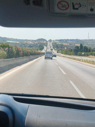 The image is a first-person perspective from inside a vehicle traveling on a highway. The road is asphalt with white lane markings. On the left side of the road, there is a concrete barrier topped with flowering pink bushes. Beyond the barrier, the landscape is hilly with olive groves or vineyards and scattered buildings. On the right side of the road, there is a metal guardrail. In front of the vehicle, a white van is traveling in the same lane. Further ahead, other vehicles are visible on the highway, which curves and ascends into the distance. The sky is clear and bright, suggesting a sunny day. The sun visor of the vehicle is partially visible at the top of the image, with warning labels for airbags. The overall scene depicts a typical highway journey through a rural, Mediterranean landscape, possibly in the region of Άγιος Παύλος, Greece, given the vegetation and the architecture.
