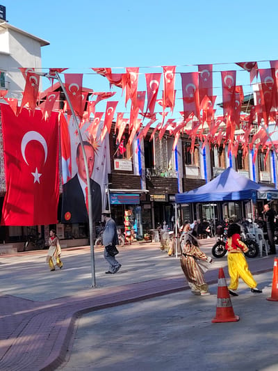 This vibrant daytime scene captures a festive street in Iznik, Turkey. The street is heavily decorated with numerous small Turkish flags strung across overhead wires, creating a canopy of red and white. A large Turkish flag, emblazoned with the white star and crescent, hangs prominently on the left side of the frame. Adjacent to it is an equally large banner featuring a portrait of Turkish President Recep Tayyip Erdoğan.

The street is bustling with activity, with several people visible. Many individuals, including children, are dressed in colorful, traditional-looking attire, suggesting a cultural event or celebration. Some are walking across the street, while others are gathered under a blue canopy tent on the right side. Buildings with multi-story facades line the street, housing various businesses; a visible sign reads "ERKARDESLER GAYRİMENKUL." Other partially visible signs include "TURKIYE" and "EKONOMi."

The sky is clear and bright blue, and strong sunlight casts distinct shadows, indicating a sunny day. Orange traffic cones are placed on the street, likely for crowd control or to mark an area. Bicycles and motorbikes are parked intermittently along the street. The overall impression is that of a public holiday or significant cultural event being celebrated.