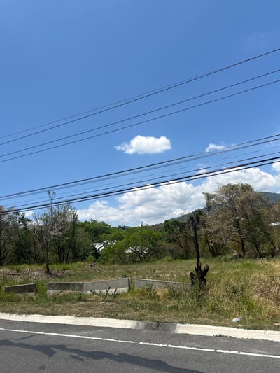 The scene is a view from the edge of a paved road in Pozos, Costa Rica (latitude: 9.948, longitude: -84.177). The road is empty, with a white line down the center. A grassy area with a few scattered trees is to the right of the road, behind a concrete barrier. The sky is blue with some scattered white clouds. There are multiple power lines running across the sky. A few trees and foliage obscure the view of any buildings or houses in the distance.