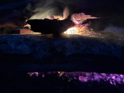 The image depicts a close-up, low-angle shot of burning logs at night. The logs are arranged horizontally, with smoke and flames rising from the top log, creating an ethereal glow. The fire's light illuminates the charred wood, revealing textures and cracks. Below the burning logs, there's a dark, indistinct surface that appears to reflect the fire's colors, with a notable purple hue in the lower portion of the frame. The overall scene is dark and atmospheric, suggesting an outdoor setting, possibly a campfire or fireplace. There is no visible indication of a specific location within the city of Ardu, Estonia. The image focuses solely on the natural phenomenon of fire and burning wood.