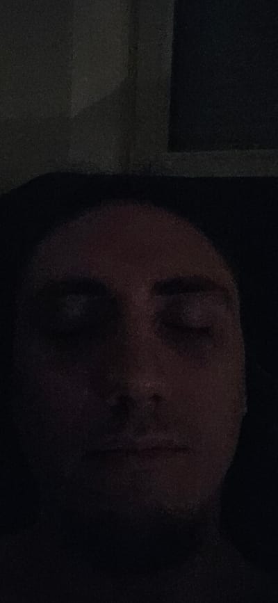 The image is a close-up, low-light photograph of a person's face. The person's eyes are closed, and their head is tilted slightly. The lighting is dim, casting shadows on the face and making details difficult to discern. The background is dark and appears to be an indoor setting. The image does not contain any information that would allow for the identification of the location within the city of Villing, France, or any specific details about the scene beyond the facial features of the subject.