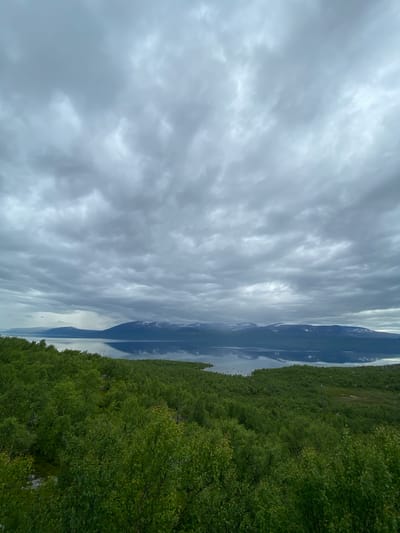 This is a photograph taken in Kiruna, Sweden, featuring a serene natural landscape. The scene is dominated by a vast, calm body of water reflecting the sky and the surrounding environment. To the right of the water, a dense, lush green forest stretches into the distance, with the foliage appearing vibrant and healthy. The sky above is filled with dramatic, heavy clouds, suggesting overcast or possibly stormy weather. The light is diffused, creating a soft and somewhat muted atmosphere. The reflection in the water is remarkably clear, mirroring the cloudy sky and the dark silhouettes of mountains or hills in the far distance. There are no people or man-made objects prominently visible, emphasizing the wild and untouched nature of the location. The overall impression is one of peaceful solitude and the raw beauty of the Arctic wilderness. The time of day is difficult to pinpoint precisely due to the overcast conditions, but it appears to be daytime.