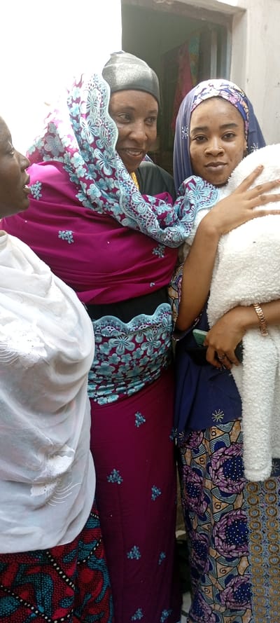 In Dan Maliki, Nigeria, three women are depicted in a close-up, vertical shot.
The woman on the left, partially visible from the chest up, wears a white sheer headscarf and a red and teal patterned garment. Her face is mostly out of frame, showing only the side of her head.
The central woman, positioned slightly behind the other two, wears a dark head covering, possibly a cap, and a magenta garment with teal and light blue floral embroidery. She has a gold-colored necklace visible. Her face is turned towards the left, with a smiling expression, and her eyes are looking in that direction.
The woman on the right, in the foreground, wears a dark blue headscarf patterned with light blue dots and a nose piercing. She holds a white, fluffy, possibly fleece-like object against her chest with both hands. Her left wrist displays a rose gold-colored watch or bracelet. A smartphone with a light-colored screen is partially visible in her left hand. She wears a patterned dress featuring blue, purple, and gold tones. Her gaze is directed towards the camera.
The background consists of a light-colored, possibly white or off-white wall on the left and a darker doorway or interior space on the right, suggesting an indoor or semi-outdoor setting. The lighting is bright, casting soft shadows.