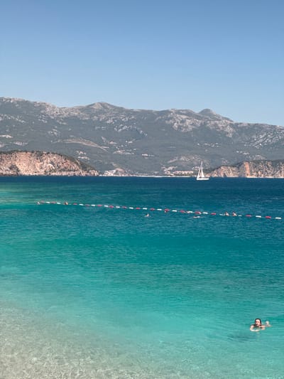 A vertical image depicts a sunlit coastal scene. The upper portion features a clear, cloudless, light blue sky. Below, a distant mountain range with dark green vegetation and exposed grey rock faces spans the horizon, with sparse residential structures visible on its lower slopes and coastline. A prominent reddish-brown and grey rocky cliff extends from the left side towards the center.
The middle ground consists of deep blue sea, where a white sailboat with a single raised main sail is positioned slightly right of center. A horizontal line of alternating red and white buoys stretches across this section, acting as a swimming demarcation. Behind this buoy line, approximately seven individuals are visible swimming, with only their heads above the water.
The foreground is dominated by exceptionally clear, vibrant turquoise water that gradually becomes lighter towards the bottom, revealing a light-colored, pebbly seabed. In the lower right quadrant, a woman swims in this clear water, facing slightly towards the viewer, with her head and upper chest visible above the surface. No specific city identifying details for Kubasi, Montenegro are discernible within the image itself.