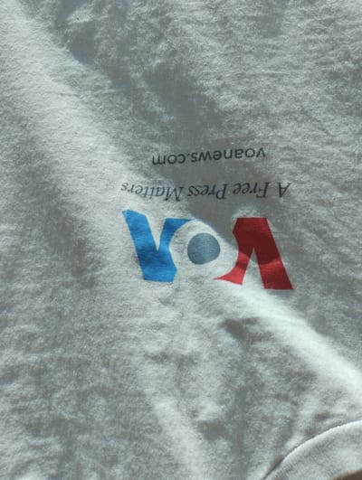 A white t-shirt displays the VOA logo in blue, gray, and red. Below the logo, the text "A Free Press Matters" is printed, followed by "voanews.com." The shirt appears slightly wrinkled, and the fabric has a textured look, possibly due to the lighting.