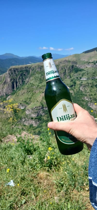 A person's hand, wearing a blue terrycloth sleeve, holds a green glass bottle of Dilijan beer. The bottle's label is predominantly white with green accents and features the text "Dilijan Beer" in green lettering. The background is a mountainous landscape with steep, green cliffs, a clear blue sky with sparse clouds, and patches of yellow wildflowers in the foreground. The location is identified as Dsegh, Armenia, a village known for its scenic natural beauty and as the birthplace of writer Hovhannes Tumanyan. The scene appears to be an outdoor setting on a sunny day.
