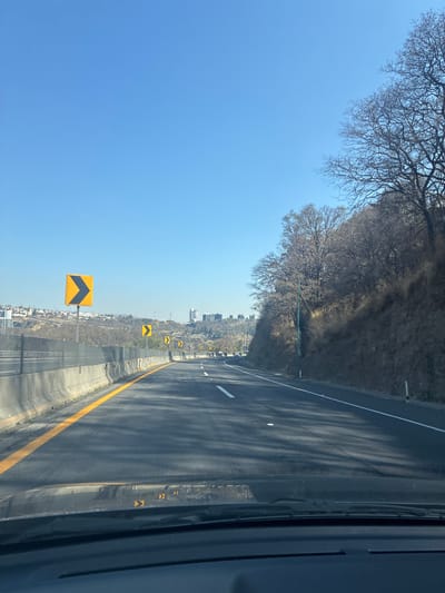 The image depicts a highway in Santiago Yancuitlalpan, Mexico. The road curves to the right and is lined with yellow reflective markers on the road's edge. There are black-and-yellow chevron signs along the side of the road to warn drivers of the curve. Buildings and greenery can be seen in the distance. The sky is blue and clear. A forested area is visible on the left side of the highway. The image appears to be taken from inside a vehicle traveling on the highway.