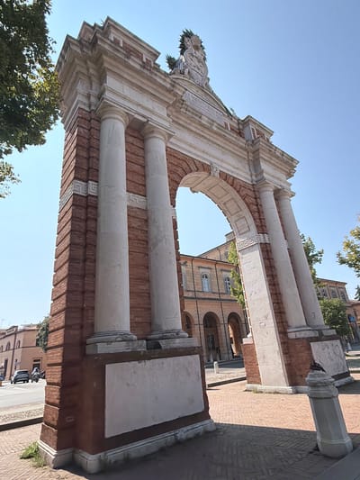 The image shows a low-angle view of the Triumphal Arch of Clement XIII in Santarcangelo di Romagna, Italy. The arch is constructed from red brick and features white marble decorative elements, including columns, cornices, and a sculpted crest at the apex. The inscription on the arch is partially visible. The sky is clear blue. In the background, a building with arched walkways and windows can be seen, along with trees and a street with a car and a scooter. The ground in front of the arch is paved with bricks.