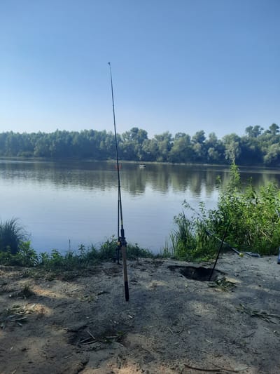 This is a photograph taken on the bank of the Desna River in the village of Pukhivka, Ukraine, on a clear, sunny day. Two fishing rods are planted in the sandy ground, ready for use. In the middle distance, a small boat with at least two occupants is visible on the calm, reflective water. The opposite bank is lined with a dense, verdant forest. The foreground is a sandy area with scattered fallen leaves. The scene conveys a peaceful, recreational setting.