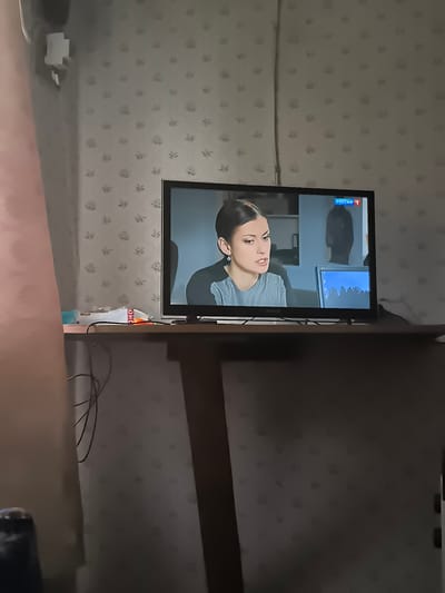 The image shows a television screen displaying a program. On the screen, a woman is visible in the foreground, looking to her left with an intense expression. She is wearing a grey top and a diamond earring. Behind her, a man's silhouette is visible, and to the right, a computer monitor displays a landscape scene with snow-covered mountains and trees. The television itself is a flat-screen model with a black bezel. A red and blue logo with the word "РОССИЯ 1" is in the top right corner of the screen.

The television is placed on a shelf or table, which is cluttered with a few items including a white package with red writing that appears to say "OHO", and a blue object. Cables are visible behind the television. The background wall has a light-colored floral wallpaper pattern. To the left of the television, a pink curtain or fabric is draped. The lighting suggests an indoor setting, likely in the evening or on a cloudy day, as the ambient light is dim. The overall mood of the image is domestic, focused on the act of watching television. There are no clear indicators of time of day or weather conditions outside of the indoor setting. The location is specified as Posadnikov Ostrov, Russia.