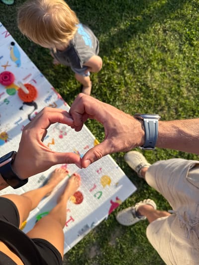 In Зажэвіцкі сельскі Савет, Belarus, a medium shot captures an overhead view of a family on a sunny day. A toddler with blonde hair, wearing a grey t-shirt, stands on a white play mat with colorful cartoon animals and shapes, on a lush green lawn. Two adults' hands are in the foreground, forming a heart shape. One adult, wearing a black watch and shorts, is barefoot, with painted red toenails. The other adult, wearing khaki shorts and light-colored crocs, has a watch with a grey band. The scene is framed by the adults' legs and feet, suggesting a casual outdoor setting, possibly a backyard or park.