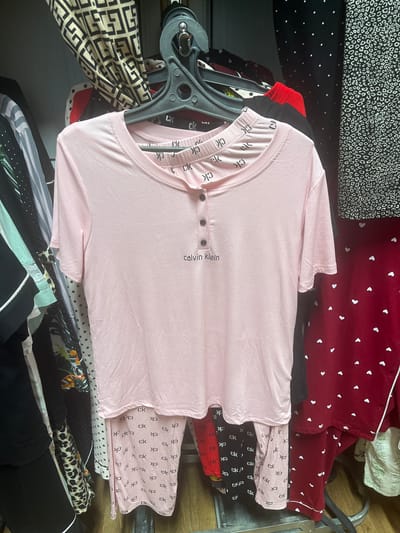 The image shows a close-up of a pink pajama set hanging on a rack in a store. The pajama top is short-sleeved with a ribbed texture and a Henley neckline with three buttons. The words "Calvin Klein" are printed in small black letters on the left side of the chest. The pajama bottoms are also pink with a ribbed texture and are covered in repeating black "ck" logos. Various other patterned garments are visible in the background, suggesting a retail environment. The lighting is somewhat dim, typical of an indoor store setting. There are no people or animals present in the image. There is no information in the image that places it in the city of Şü, Kazakhstan.
