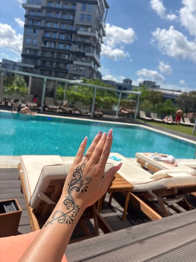 A hand with a henna tattoo is in the foreground, with a swimming pool and lounge chairs behind it. In the background, a modern high-rise building stands under a clear blue sky with scattered clouds, suggesting an urban setting in Kâğıthane, Turkey. Several people are visible near the pool, some lounging on chairs and others in the water. The scene appears to be a resort or hotel with outdoor amenities. The lighting suggests it is daytime, with bright sunlight casting shadows.