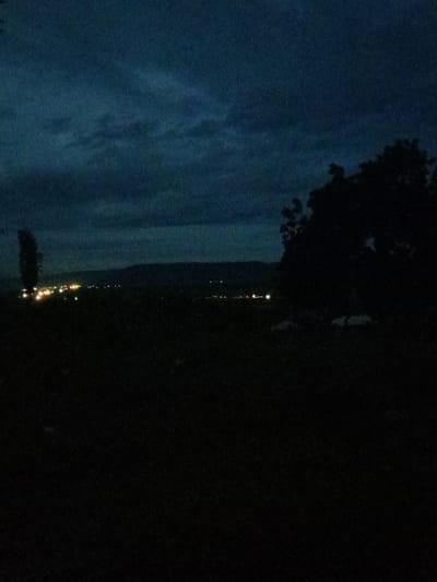 A dark landscape image depicts a night scene with city lights in the distance, blurred by the atmospheric conditions. The foreground is deep black, suggesting a field or unlit area. The sky is a blend of dark blue and cloudy patches, contributing to the overall dim and somber atmosphere. Silhouetted trees are visible on the right side of the frame, adding depth and framing the distant cityscape.