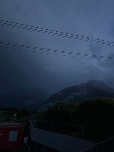 The image depicts a dark, moody landscape at dusk or during a storm, likely in or near Hasliberg Reuti, Switzerland. The sky is filled with heavy, dark clouds, casting a deep blue-grey hue over the scene. Several power lines stretch horizontally across the upper portion of the image. In the middle ground, a large, imposing mountain range is visible, with its peaks partially obscured by the low light and clouds. Some snow or lighter rock formations are visible on the mountain slopes. In the foreground, a dense hedge or thicket of trees runs horizontally, separating the viewer from the background. To the left, a red shipping container with a white window is partially visible, along with some other structures and equipment that appear to be on a construction site or industrial area. Further back and to the left, a small house with illuminated windows is nestled amongst trees on a green hillside. The overall atmosphere is somber and dramatic, suggesting inclement weather or the late hour.