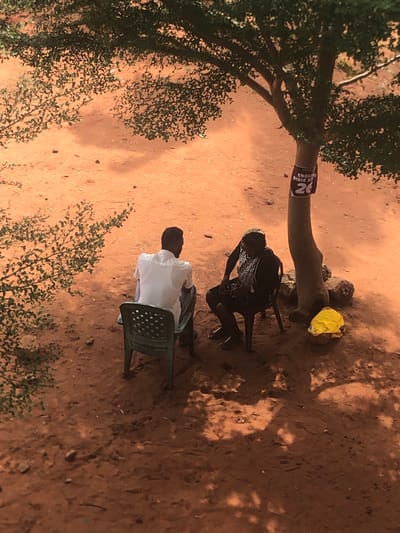 The scene is an outdoor setting in Nteje, Nigeria. The ground is reddish-brown soil, partially shaded by trees. Two people are present. One individual is seated in a green plastic chair, wearing a white shirt. Another individual is seated in a black chair, wearing dark clothing with a light-colored pattern on the upper garment. A yellow cloth lies near the base of a tree. The tree has a sign affixed to it, partially obscured, displaying the text "ENGLISH BIBLE ST".