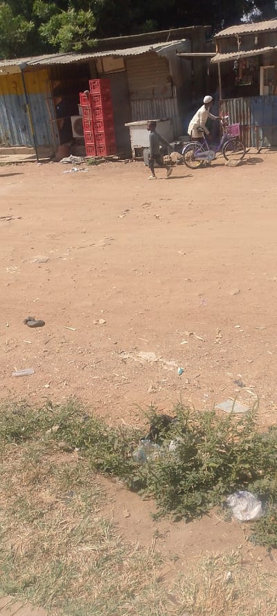 This image depicts a daytime scene on an unpaved, reddish-brown dirt thoroughfare in Danbare, Nigeria.

**People:**
1.  A young male, likely a child or adolescent, is in the mid-ground, moving from left to right across the dirt path. He is wearing dark trousers, a long-sleeved dark shirt, and light-colored shoes. His left leg is extended forward.
2.  An adult male is standing in the mid-ground, to the right of the young male. He is wearing a light-colored, long-sleeved garment, possibly a traditional kaftan, and a white cap. He is positioned next to a bicycle.

**Subjects/Objects:**
*   **Structures:** In the background, there are multiple single-story commercial structures made primarily of corrugated metal sheets with pitched corrugated metal roofs. The leftmost structure exhibits sections painted yellow and blue.
*   **Crates:** Outside the leftmost structure, a vertical stack of approximately eight red plastic beverage crates, consistent with Coca-Cola branding, is visible.
*   **Appliance:** A white cooler or similar appliance is situated on the ground near the stack of red crates.
*   **Bicycle:** A purple bicycle with a pink basket attached to its handlebars is stationary next to the adult male.
*   **Ground:** The immediate and mid-ground consist of unpaved, uneven reddish-brown dirt. Scattered debris, including plastic bags and fragments of material, is present across the dirt surface.
*   **Vegetation:** Sparse, dry grass and low-lying green shrubs are visible in the foreground along the bottom edge of the image. Further back, above the commercial structures, a line of green trees is present.

**Actions/Interactions:**
*   The young male is captured in mid-stride, suggesting movement.
*   The adult male is standing next to his bicycle, appearing stationary.

**Environment:**
The scene is brightly lit by natural sunlight, indicating an outdoor, daytime setting. The environment appears to be a local commercial area or street in a semi-urban or rural context.