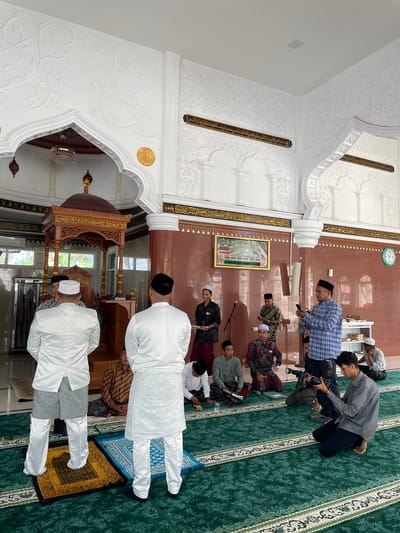 The image captures an indoor scene within Mesjid Reubee, Indonesia, depicting a gathering of men, likely for a religious event or prayer session. The setting features ornate white archways and columns with intricate carvings, contrasting with brown-tiled walls. The floor is covered with a green patterned carpet, and individual prayer mats (one blue, one golden) are visible in the foreground.
Several men are present, dressed in a mix of traditional Islamic attire and more casual clothing. Two men in the immediate foreground wear white long-sleeved tunics over white trousers and black or white kufis/songkoks, standing with their backs to the viewer. Further into the room, other men are seated on the green carpet, some wearing checkered shirts, traditional patterned sarongs, or plain garments, often topped with kufis. One man in a gray striped shirt is seen kneeling and operating a camera with a large lens, suggesting documentation of the event. Another man is seated, holding what appears to be a religious book on a small wooden stand.
Architectural details include a richly carved wooden minbar or pulpit with a domed top, visible towards the left side of the frame. Golden-colored strips of Arabic calligraphy adorn the white archways and parts of the brown walls. A framed picture, possibly depicting a religious site or text, hangs on the brown wall, and a circular wall clock indicates the time is approximately 12:45.
Visible text includes the word "MASJID" and various lines of Arabic script from the provided OCR, such as "الات المواليوم التالي يتخوف من الاقواي سي بحريه واستغفروان كان توابا النصر". The atmosphere appears calm and focused, characteristic of a religious gathering.