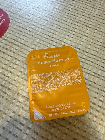 A close-up shot displays a packet of Chick-fil-A Honey Mustard sauce resting on a textured surface. The packet is predominantly yellow with the Chick-fil-A logo and "Honey Mustard Sauce" printed on its front. A list of ingredients is detailed along the side, including water, honey, sugar, distilled vinegar, and modified corn starch, among others. The bottom of the packet indicates it is packed for Chick-fil-A, Inc. in Atlanta, GA 30349, and states the net weight as 1 oz (28g). Part of a round item with the word "Order" is visible on the bottom left corner. The surface beneath the packet is a light beige with a repetitive textured pattern. The photograph offers a detailed view of the product packaging. It is not possible to verify the location is Stone Mountain.