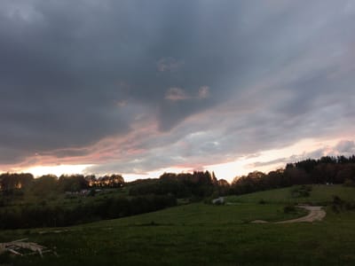 The image shows a landscape at dusk or dawn, likely in a rural area. A large expanse of grass and a dirt road is visible in the foreground. The middle ground includes a hill with scattered trees. In the distance, a line of trees and possibly a small building are silhouetted against the sky. The sky is filled with dark, heavy clouds, with hints of pink and orange suggesting the sun is setting or rising. There is no evidence to suggest the exact location in Bogoslov, Bulgaria.