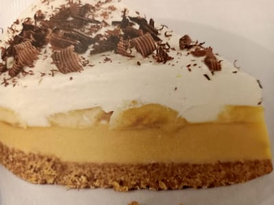 The image is a close-up, overhead view of a slice of banoffee pie on a white plate. The pie has a crumbly, brown crust, a thick layer of yellow filling, and is topped with white whipped cream. The whipped cream is garnished with numerous dark chocolate shavings. The lighting is bright, and the focus is sharp on the pie. There are no people or any discernible features of Rumaithiya, Kuwait, visible in the image.