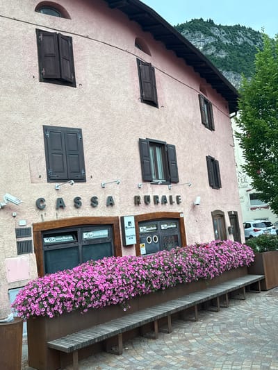 This is a medium-shot, eye-level, outdoor photograph of a building facade in Fai della Paganella, Italy. The building is painted a light pink and has dark brown shutters on its windows. A sign above the ground floor windows reads "CASSA RURALE." The ground floor features two windows with signs that say "La Cassa Rurale" and one with "Sportello ATM." A row of pink flowers cascades over a planter box in front of the windows, and a wooden bench sits in front of the planter. To the right of the building, a white car is parked. A lush green tree stands on the right side of the frame, and a mountainous landscape is visible in the background. The sky is overcast.