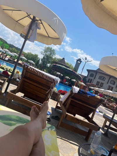 The image depicts a sunny day at a resort or spa in Gorna Malina, Bulgaria, with a clear blue sky and white, puffy clouds. The foreground features a person's legs and feet resting on a yellow and green patterned lounger, with a second person's feet visible near the lounger. Wooden lounge chairs, adorned with white towels, are positioned around a swimming pool. A large white umbrella casts shade over one of the lounge chairs, with a blue shirt and some papers hanging from its pole. In the background, the swimming pool is visible with several people relaxing. Further back, a stone building with a tower-like structure and red-tiled roofs, surrounded by greenery and flowers, is prominent. The overall scene conveys a relaxed atmosphere of leisure and vacation.