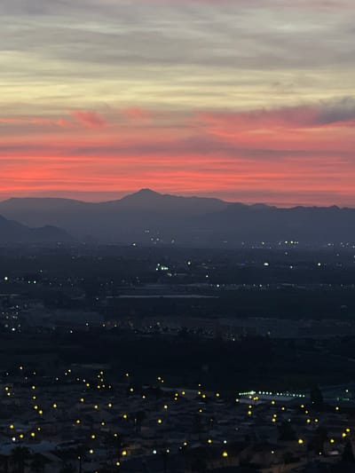Here is a detailed description of the image:
The image shows a night view from an elevated location in Rojales, Spain. The sky features a vibrant sunset with colors ranging from pale grey and yellow on the left to vivid orange and red on the right. The silhouette of a mountain range is visible against the twilight sky. Below the mountains lies a sprawling urban area dotted with numerous lights, indicating residential or commercial buildings. The scene suggests that the image was taken during dusk or early evening.