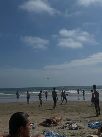 The scene is on a beach in Montanita, Ecuador, located at latitude -1.825 and longitude -80.755. The photo was taken on a sunny day, with a blue sky and white fluffy clouds. The beach is sandy, with a calm, blue ocean in the background. There are several people on the beach, with some playing a game of soccer with a yellow ball. Others are sitting on towels, watching the game. A woman lays on a beach towel, face down, in the foreground. There are a few towels and blankets on the ground near her. A man is partially visible on the left side of the photo, sitting down and looking towards the camera. There are small waves breaking on the shore. There is a large, dark blue blanket, folded, on the sand in the lower right side of the photo, near a blue beach bag.
