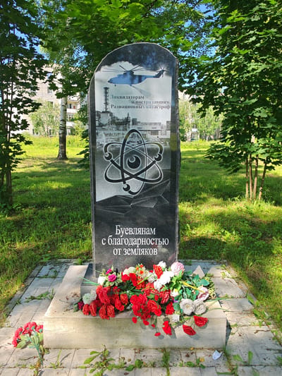 This media file shows a monument dedicated to the liquidators and victims of radiation disasters. The monument, made of black granite, is carved with images of a helicopter, industrial structures resembling a power plant, and an atomic symbol. Below the imagery, Russian text reads, "To the liquidators and victims of radiation disasters with gratitude from the people." The monument stands in a grassy area with trees and residential buildings visible in the background, suggesting an outdoor setting, likely in a town or city. Artificial flowers, predominantly red and white, are placed at the base of the monument, indicating a memorial or a site of remembrance. The lighting suggests it is daytime, with sunlight casting shadows, and the trees are full of green leaves, indicating a temperate season. The scene evokes a somber and respectful atmosphere, honoring those affected by nuclear accidents. The location context provided is Buy, Russia.