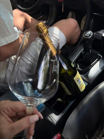 The image shows a close-up view from inside a car, looking towards the center console and the passenger's leg. A person's bare leg, clad in white shorts, is visible. In the foreground, a hand with manicured nails is holding a champagne flute, partially filled with a bubbly liquid. The flute is angled towards a bottle of Cava, which is lying horizontally in the car's console. The Cava bottle has a yellow label with text including "Cava", "Gran Reserva", and a QR code. Next to the Cava bottle, another bottle, possibly also Cava, is visible. The car's gear shift is prominent, indicating a manual transmission. The lighting suggests it is daytime, with sunlight streaming into the car, creating reflections and highlights. The overall scene evokes a sense of relaxed celebration or indulgence. No specific location cues beyond "Unknown, Spain" are discernible.