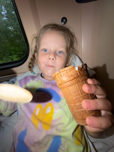 A young girl with blonde hair in pigtails is holding an ice cream cone and a spoon. She has ice cream on her face. She is wearing a tie-dye shirt with a yellow graphic and the word "smile". The girl is seated inside a vehicle, possibly a train, as indicated by the window showing trees and greenery outside. The interior of the vehicle appears to be beige and brown.