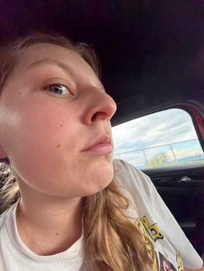 A close-up, angled shot of a young woman's face, appearing to be taken from inside a vehicle. The woman has fair skin, light brown hair, and blue eyes. She is wearing a white t-shirt. The background shows a window with a glimpse of a landscape, including a blue sky and what seems to be a distant body of water. It is not possible to determine whether the location is in the city of Chokoba, Bulgaria.
