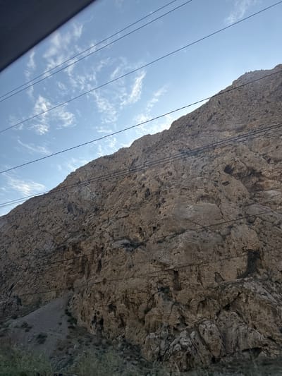 The image shows a rocky mountain slope under a clear blue sky with scattered white clouds. Several power lines run horizontally across the frame, obscuring parts of the mountain. The mountain is rugged and appears to be made of large, uneven rocks with some sparse vegetation at its base. The lighting suggests it is daytime. The scene is likely located near Kadamjay, Kyrgyzstan, based on the request, but the image itself does not contain any explicit indicators of this location. There are no people or any discernible actions or interactions depicted.