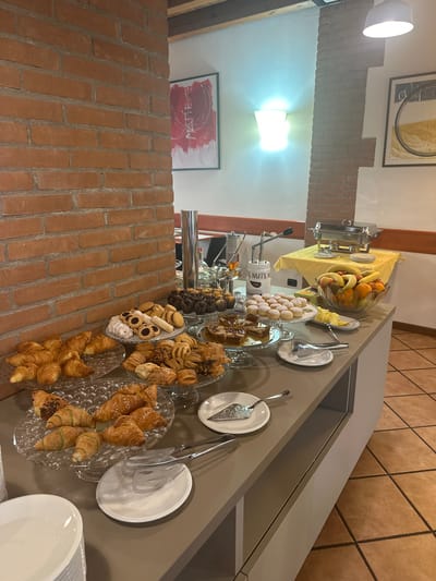 This image shows a breakfast buffet in Villafranca di Verona, Italy. The buffet is set up on a long grey table.

On the left, a brick wall provides a rustic backdrop. A food warmer, likely for keeping dishes hot, is visible at the rear of the table. A variety of pastries, including croissants, cookies, and other baked goods, are displayed on glass platters. Fruits such as bananas, oranges, and kiwis are piled in a glass bowl. Serving utensils, including tongs and spoons, are present next to the dishes. In the background, artwork adorns the wall. The floor is tiled.