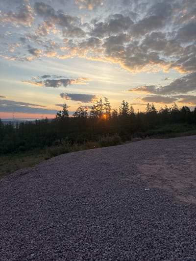 The image depicts a scenic landscape at sunset, likely in the city of Serebryany Bor, Russia, given the implied context. The foreground is dominated by a large pile of dark gray gravel, with a patch of lighter brown dirt to the right. Beyond the gravel, a dense line of evergreen trees stretches across the midground, obscuring much of the land behind them. The setting sun is visible through a gap in the trees, casting a warm orange glow and creating a starburst effect. In the distance, beyond the treeline, a body of water can be seen, with two industrial smokestacks emitting plumes of smoke on the horizon. The sky above is filled with scattered clouds, tinged with shades of orange, pink, and gray from the sunset. The overall atmosphere is tranquil and natural, with a subtle hint of industrial presence in the distance.