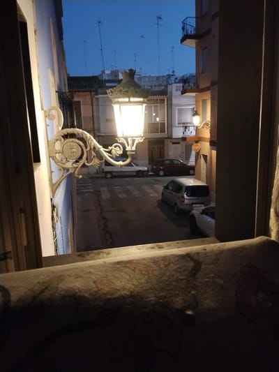 The image, taken at dusk from an elevated position looking down and across a narrow street in Sueca, Spain, captures a residential scene illuminated by artificial lights. A decorative street lamp, ornate and white, is prominently featured in the foreground, extending from a building on the left. It casts a bright, warm glow.
Below, the street is paved and marked with a pedestrian crossing. Several vehicles are parked or driving on the street. A light-colored van is positioned centrally in the mid-ground, and a dark red sedan is visible to its left, parked parallel to the curb. A lighter-colored car is partially visible further down the street. A white convertible is parked on the right side of the street, partially obscured by a building. A small, round STOP sign is affixed to the wall of a building on the right.
The architecture of the buildings lining the street suggests a European urban setting. Balconies with railings are visible on the multi-story buildings. The sky above is a deep blue, indicating twilight. The image appears to be taken through a window or from a balcony, with a portion of a patterned, dark fabric in the extreme foreground, suggesting an indoor vantage point looking outwards.