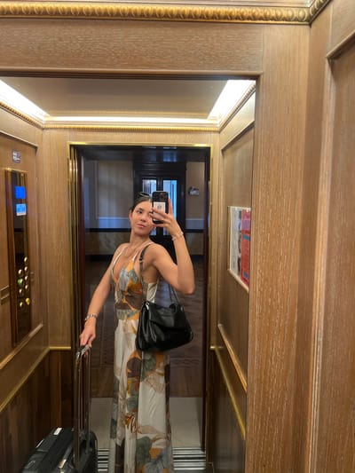 A woman stands inside an elevator, capturing her reflection in the mirror. She holds a smartphone, wearing a printed dress, and a black shoulder bag. The elevator features wood paneling, a set of buttons, and a handle for passengers. A suitcase is visible near her feet. The elevator is well-lit, with a golden frame around the mirror.