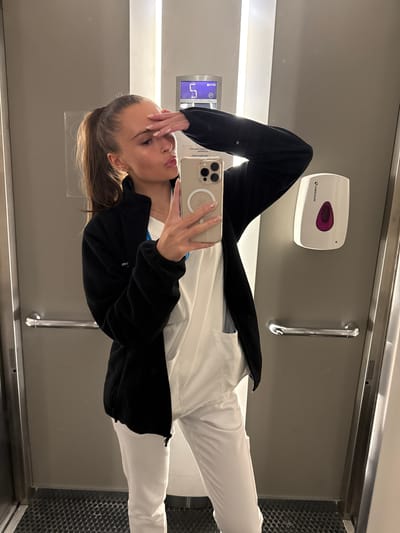 The image shows a young woman with her hair in a ponytail, wearing a black fleece jacket over a white uniform and white pants. She is holding a smartphone and taking a selfie in a mirror. Her right hand is on her forehead. Behind her, there is a hand sanitizer dispenser mounted on a wall. The environment appears to be a utilitarian space, possibly a restroom or changing room, with metallic doors and a textured floor. The lighting is bright, coming from overhead fixtures. The image does not contain any information that would allow for determination of its location in Harstad, Norway.