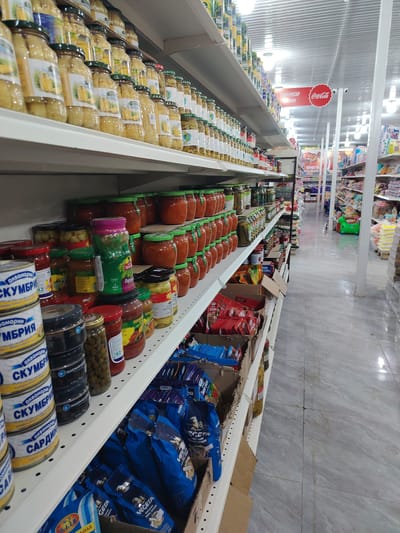 This image depicts the interior of a grocery store aisle, likely in Bosteri, Kyrgyzstan, given the Cyrillic text on product labels. The shelves are stocked with a variety of canned goods, jars of preserved food, and packaged dry goods. On the left, stacked cans with labels including "СКУМБРИ" (mackerel) and "САРДИНА" (sardines) are visible. Further along the shelf are jars of pickled vegetables and sauces, with labels such as "Горош" (peas) and "ОСТРЫТ" (spicy). In the foreground, on a lower shelf, blue and red packaged products, including some with the brand name "VEGETA," are displayed. The aisle extends into the background, showing more shelves of products and a bright, possibly artificial, lighting overhead. A red sign with "Coca-Cola" is visible in the distance. The scene suggests a typical grocery store environment, with no people visible. The overall impression is one of abundance and variety in the available foodstuffs.