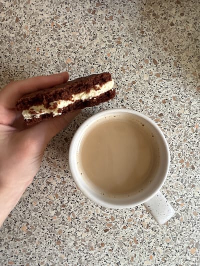 An overhead shot shows a hand holding a chocolate cookie sandwich with a cream filling. Beside the cookie is a white mug filled with a light brown beverage, likely coffee with milk, on a speckled gray countertop. The image was taken in the city of Wörth am Main, Germany.