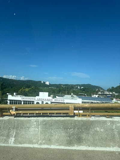 The image displays a daytime scene in Puerto Marqués, Mexico, featuring a prominent building, lush hills, and a clear blue sky. In the immediate foreground, a weathered grey concrete barrier, possibly part of a bridge or highway, is visible. Above this barrier, a yellow metal guardrail, showing signs of wear and rust, stretches horizontally across the frame.
The central subject is a white, multi-story building identified by the large dark letters "INSTITUTO LEONARDO BRAVO" prominently displayed on its facade. The building features multiple rectangular windows with dark panes. To the right of this main structure, another section of a roof is visible, equipped with multiple solar panels.
Behind the building, the landscape consists of densely vegetated green hills, characteristic of a tropical environment, with numerous trees including palm species. Scattered across these hillsides are several white, modern-style buildings, suggesting residential or resort development.
The sky above is a clear, deep blue, with a few small, wispy white clouds present. A distinct, faint white crescent moon is visible in the upper-left portion of the sky, along with a smaller bright speck nearby, possibly a lens flare. No people, specific actions, or interactions are discernible within the scene.