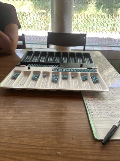 The image depicts a close-up view of a backgammon board and a notebook on a wooden table. The backgammon board is partially filled with light blue checkers. A black part of the board is visible on the left, containing rows of checkers, and a white tray with compartments holds more checkers, some arranged in groups within the compartments. Numbers are visible on some of the compartments in the white tray, including "6", "7", "8", and "9". To the right of the backgammon board, a checkered notebook is open, filled with handwritten numbers and some Cyrillic text. A black pen rests on the notebook. In the background, a window with patterned curtains allows light to filter through, suggesting an indoor setting. A person's arm and shoulder, wearing a dark red shirt, are visible on the far left of the image, indicating a person is present, likely playing the game or observing. The overall scene suggests a casual game of backgammon is in progress, with scorekeeping. The location is identified as Гүмкана, Kyrgyzstan.