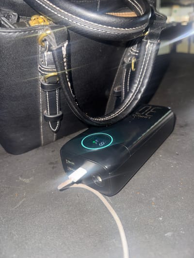 An overhead shot captures a black leather handbag and a black power bank. The handbag, with gold hardware and white stitching, is partially visible in the upper left quadrant of the image. A black USB-C cable is plugged into the power bank, which displays a blue circular indicator with a percentage reading. The scene is set on a dark, textured surface, possibly a table or floor. The lighting suggests an indoor environment, possibly a commercial or domestic setting within Abuja, Nigeria.